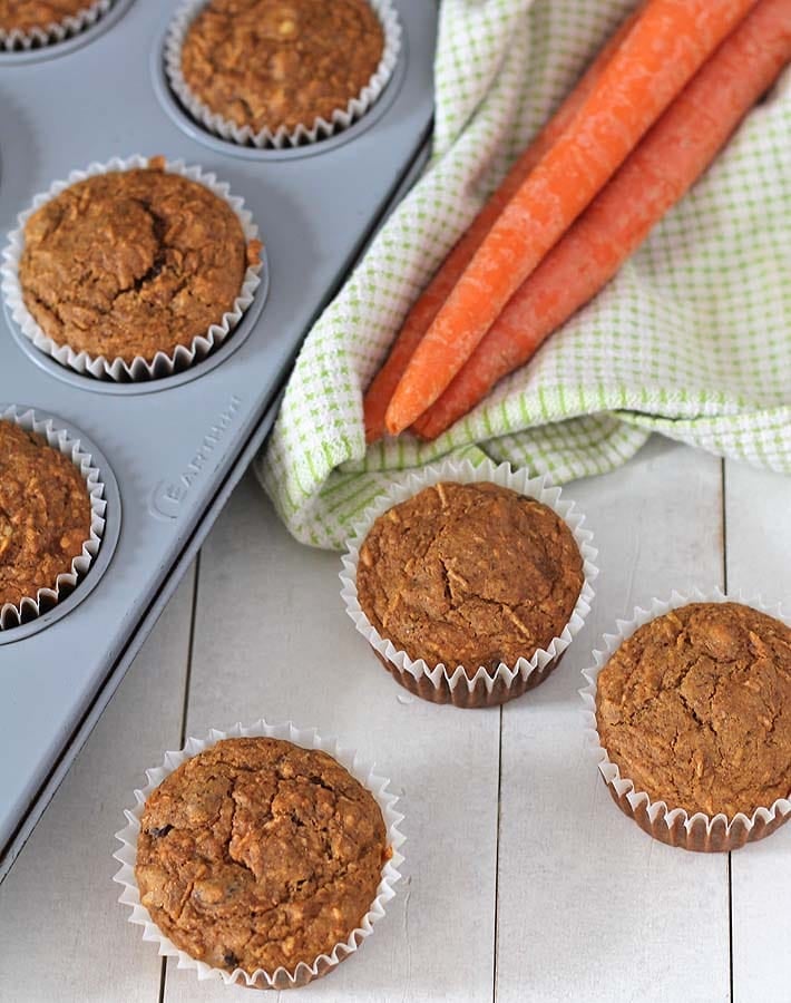 Vegan carrot muffins on a white wooden table, more muffins in a pan sit to the left.