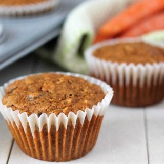 Three vegan carrot muffins sitting on a white wood table, a pan of muffins sits to the left and a green kitchen cloth with three fresh carrots on top of it sits in the background.