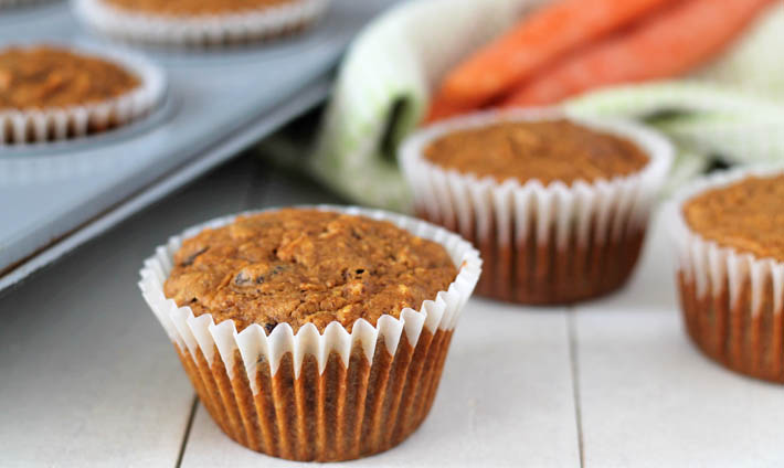 Three vegan carrot muffins sitting on a white wood table, a pan of muffins sits to the left and a green kitchen cloth with three fresh carrots on top of it sits in the background.