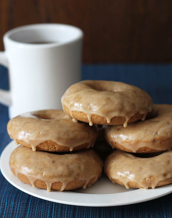 Vegan Gluten Free Maple Doughnuts stacked on top of each other on a white plate, a cup of coffee sits in the background on the left.