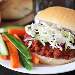 Lentil Sloppy Joes sandwich on a white plate, sandwich is topped with chopped cabbage, carrot and cucumber sticks also sit on the plate to the left.