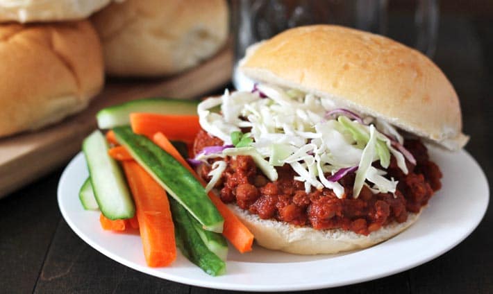Lentil Sloppy Joes sandwich on a white plate, sandwich is topped with chopped cabbage, carrot and cucumber sticks also sit on the plate to the left.