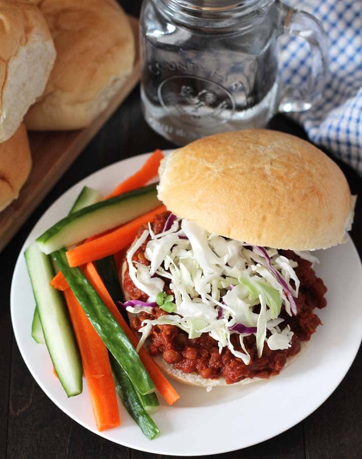 Overhead shot of a lentil sloppy joes sandwich on a white plate, sandwich is topped with chopped cabbage, plate also has cucumber and carrot sticks on it, a glass of water sits behind the plate.