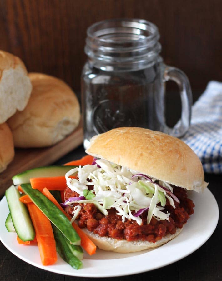 Lentil Sloppy Joes sandwich on a white plate, carrot and cucumber sticks sit to the left on the plate, a glass of water sits behind the plate and a cutting board with sandwich buns sits to the left.