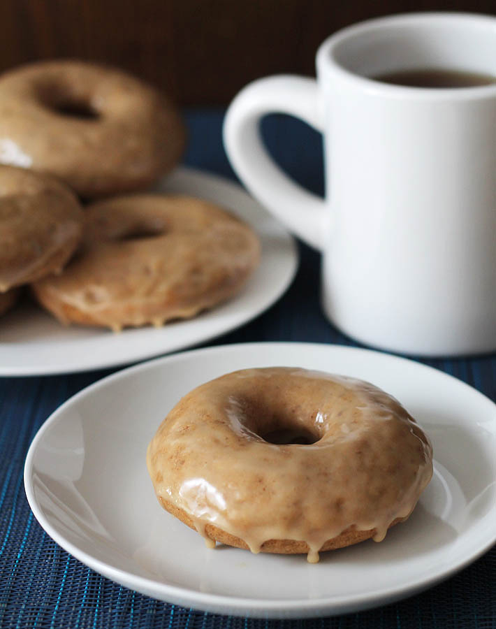 A plate of Vegan Gluten Free Maple Doughnuts sits in the background, in the foreground, one doughnut sits on a small white plate, a cup of black coffee is on the right in the background.