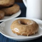 A maple doughnut on a small white plate, a white cup sits off to the right of the plate.