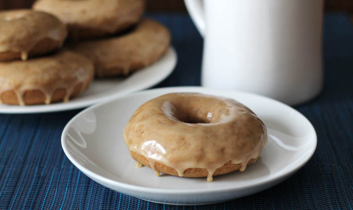 A plate of Vegan Gluten Free Maple Doughnuts sits on a blue place mat in the background on the left, in the foreground, one doughnut sits on a small white plate, a white cup sits off to the right in the background.