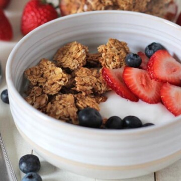 A bowl with peanut butter granola, yogurt, fresh strawberries and blueberries sitting on a white wooden table.