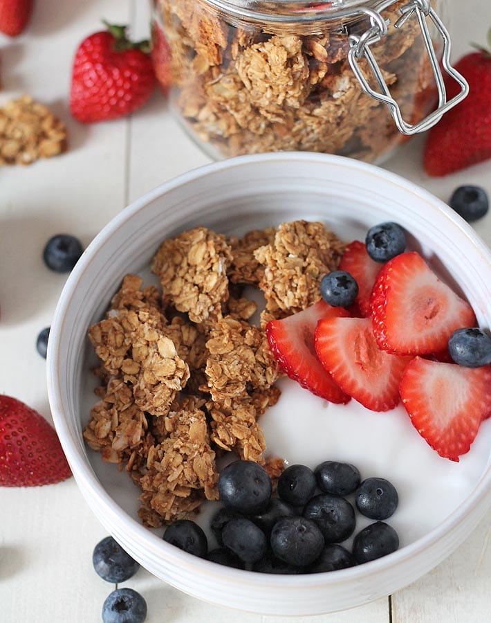 Overhead shot of peanut butter granola in a bowl of yogurt and fresh strawberries and blueberries, a jar of granola sits behind the bowl and strawberries and blueberries sits on the side of the bowl.