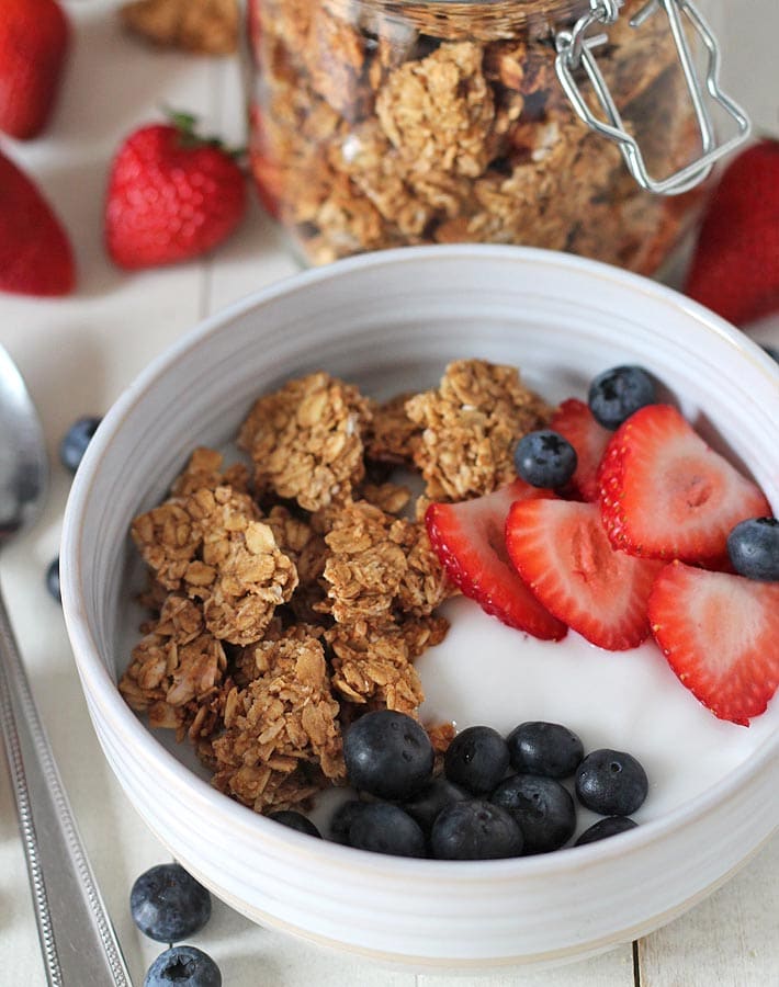 Peanut butter granola in a bowl of yogurt and fresh strawberries and blueberries on a white wooden table, a jar of granola sits behind the bowl, strawberries and blueberries sits on the side of the bowl.
