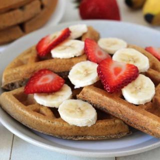 Vegan Banana Waffles on a white table in a white plate, waffles are garnished with sliced bananas and sliced strawberries.