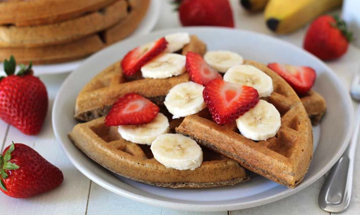 Vegan Banana Waffles on a white table in a white plate, waffles are garnished with sliced bananas and sliced strawberries.