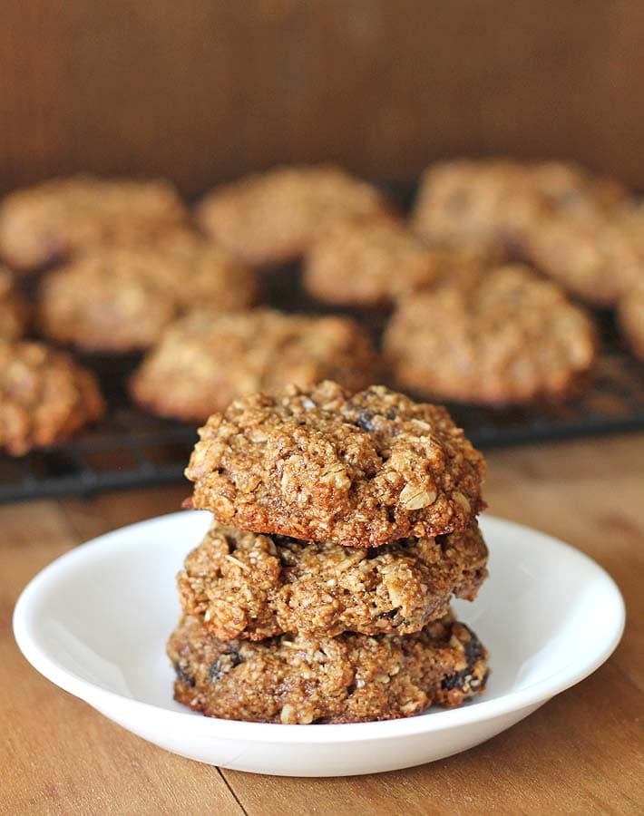 Three raisin cookies stacked on a small white plate, more cookies sit in the background on a cooling rack.