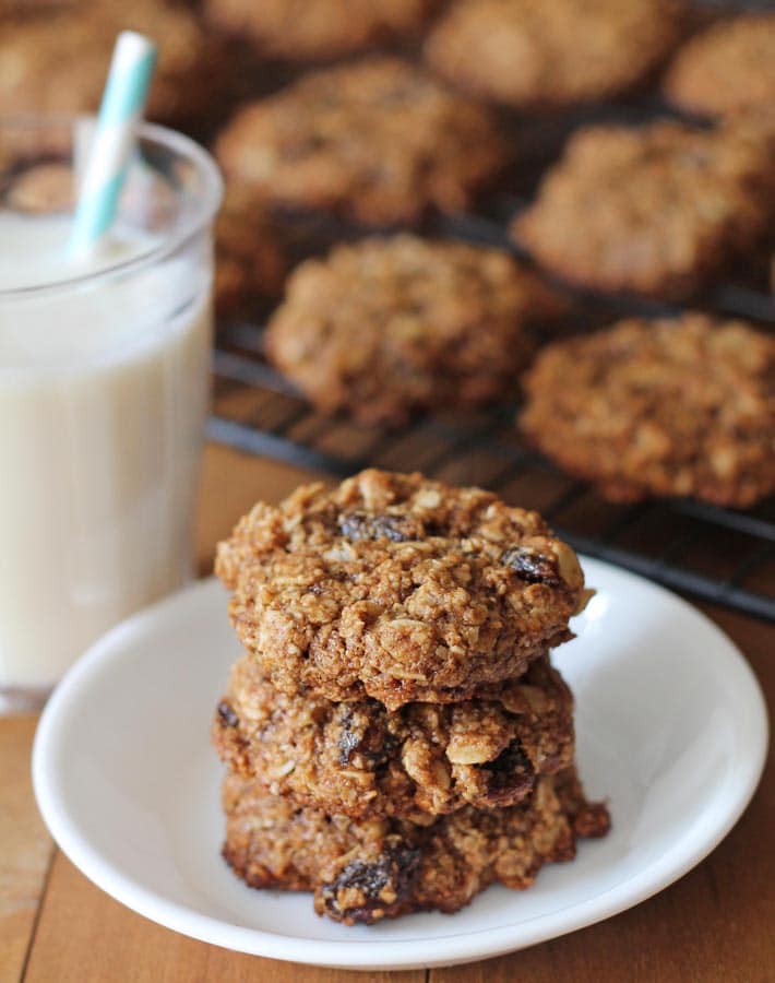 Three vegan oatmeal raisin cookies stacked on top of each other, a glass of milk sits to the left.