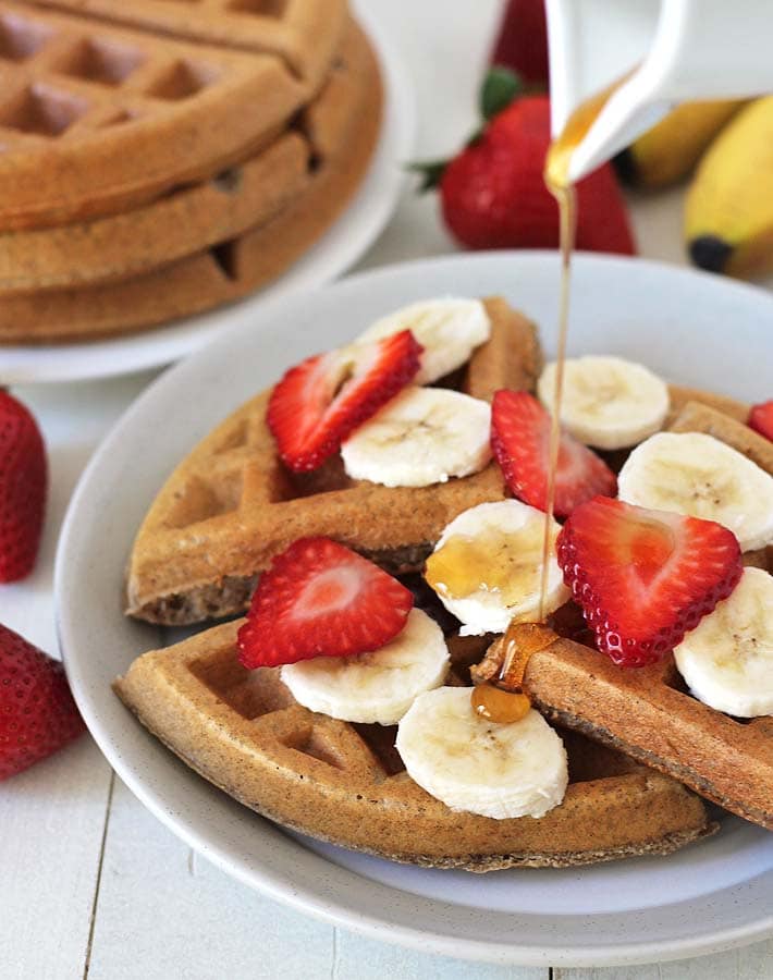 Maple syrup being poured onto gluten-free vegan banana waffles, waffles have sliced bananas and strawberries on top.