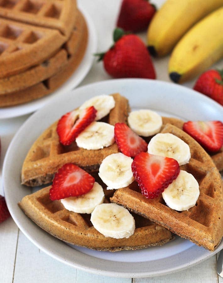 A plate of banana waffles on a white plate, bananas, strawberries, and a small cup of maple syrup sits behind the plate.