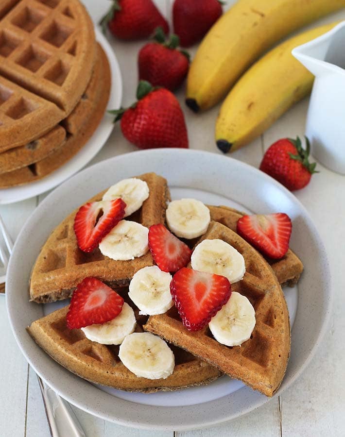 A plate of vegan gluten free banana waffles with slices of fresh strawberries and bananas on top.