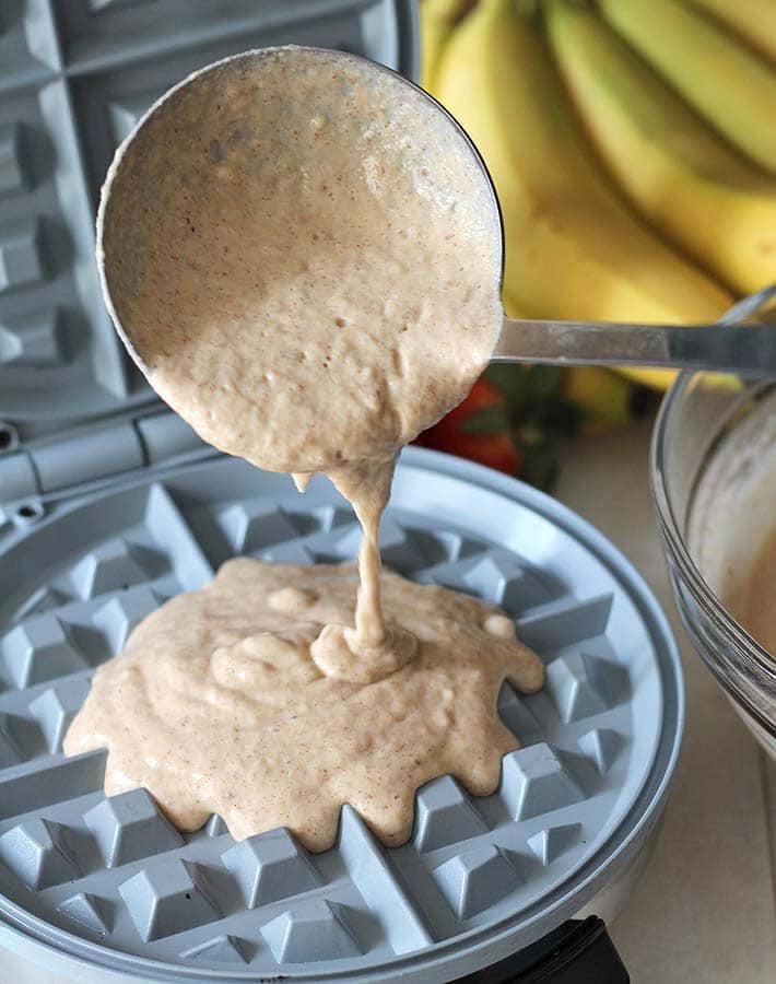 The batter for Gluten-free Vegan Banana Waffles being poured from a stainless steel ladle into a waffle maker.