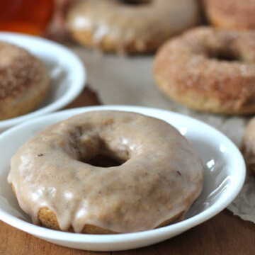 Chai-Spiced Baked Vegan Doughnuts on a wood table, two doughnuts are on white plates and the rest are on parchment paper.