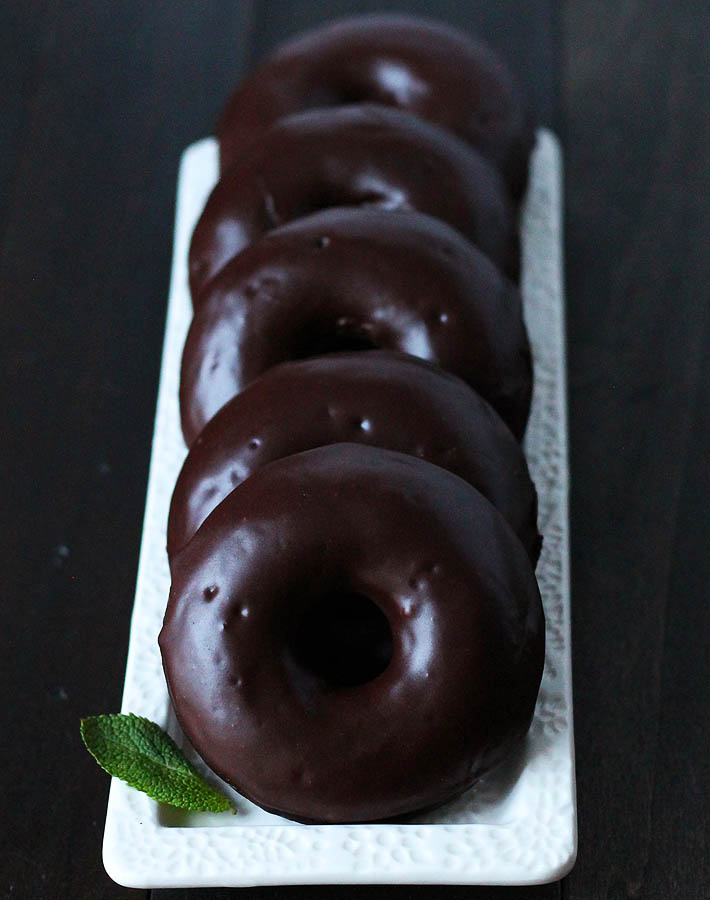 A row of five Vegan Gluten Free Chocolate Mint Doughnuts sitting on a white rectangle plate with a mint leaf to the left of the first doughnut in the row.
