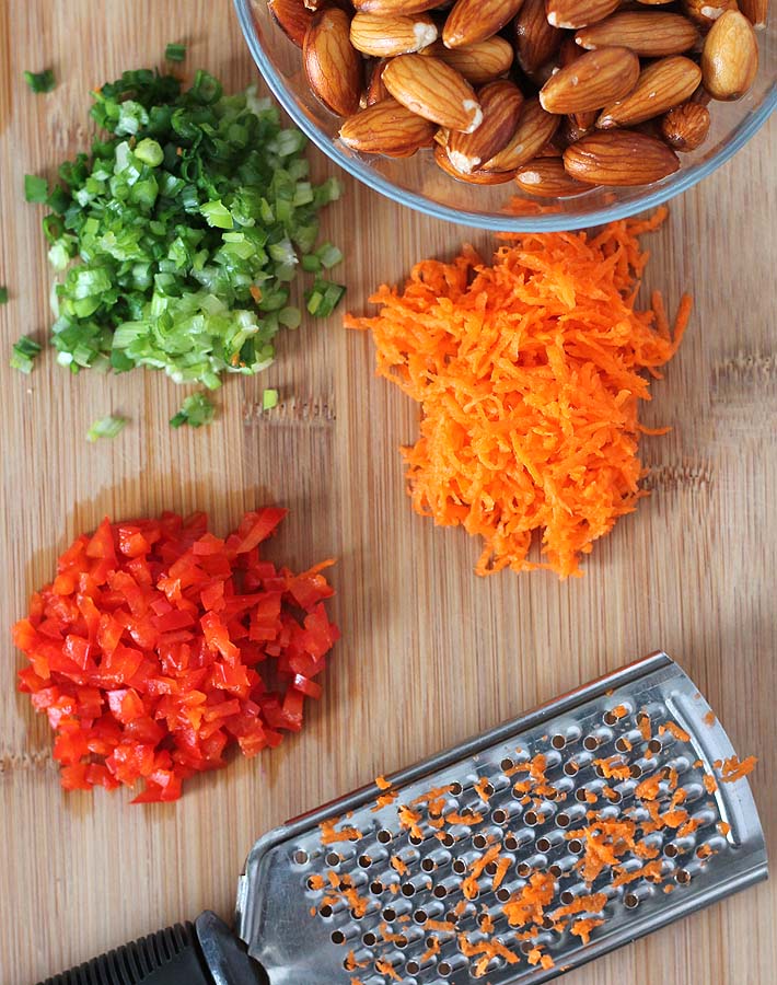 An overhead view of some of the ingredients needed to make a Vegan Vegetable Almond Cheese Ball sitting on a bamboo cutting board, ingredients show are almonds, chopped red pepper, chopped green onion and shredded carrots.