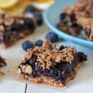 Blueberry Oatmeal Bars on a white table with fresh blueberries and lemons behind them.
