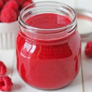 Easy Raspberry Sauce in a mason jar, jar is sitting on a white table and is surrounded by fresh raspberries.