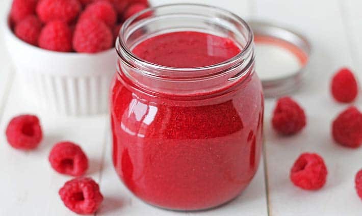 Easy Raspberry Sauce in a mason jar, jar is sitting on a white table and is surrounded by fresh raspberries.