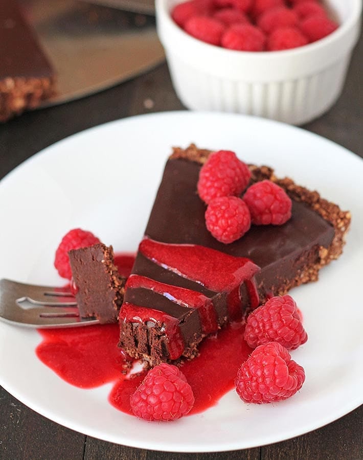 A slice of a vegan chocolate tart on a white plate with raspberry sauce and fresh raspberries on top of it.