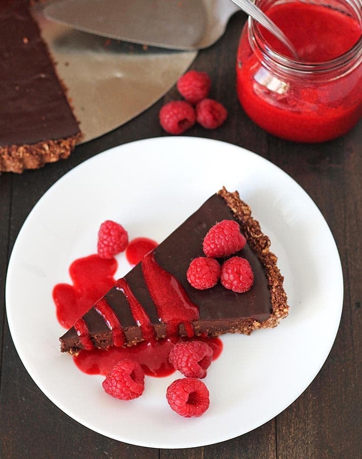 Overhead shot of a slice of vegan chocolate tart on a white plate, a jar of raspberry sauce sits behind the plate.