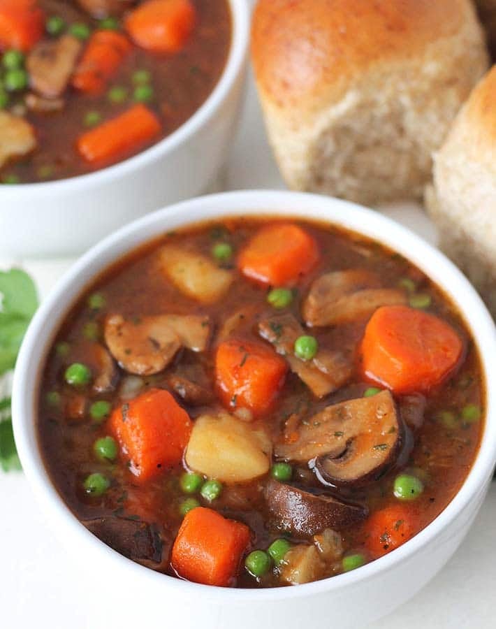 Two bowls of mushroom stew on a white table, a plate of whole wheat rolls sits in the background.