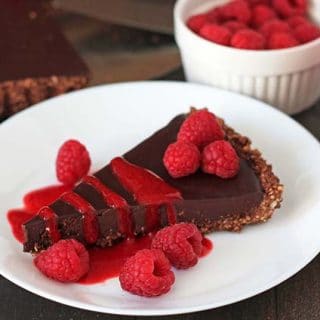 A close up shot of a slice of vegan chocolate tart on a plate, slice is garnished with raspberry sauce and fresh berries.