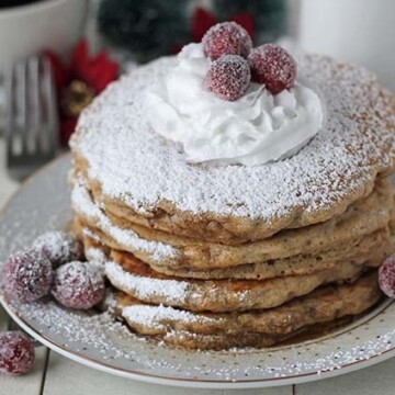 A stack of four vegan eggnog pancakes on a plate, pancakes are topped with coconut whipped cream and sugared cranberries.