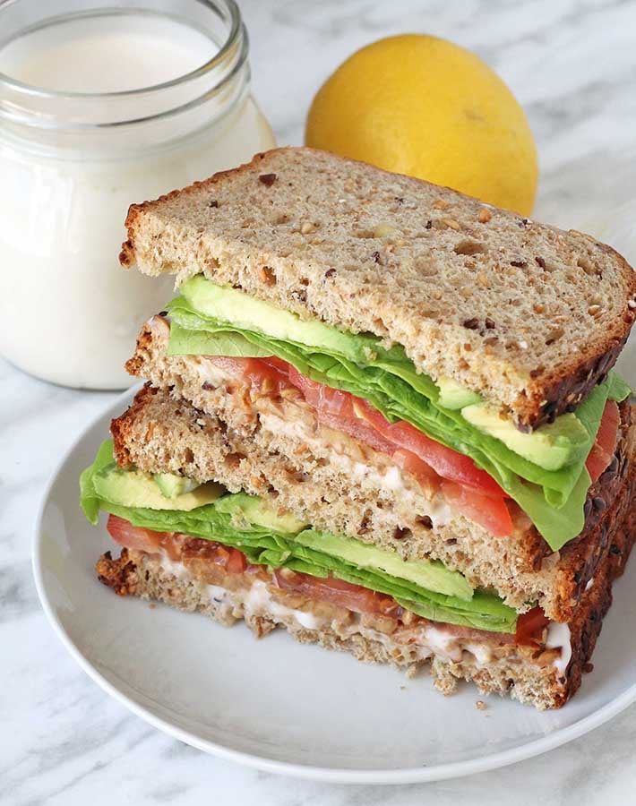 A sandwich with vegan aioli, jar of aioli and a fresh lemon sits inthe background.