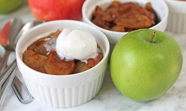 A small white bowl with baked cinnamon apple slices topped with ice cream, a green apple sits to the right of the bowl.