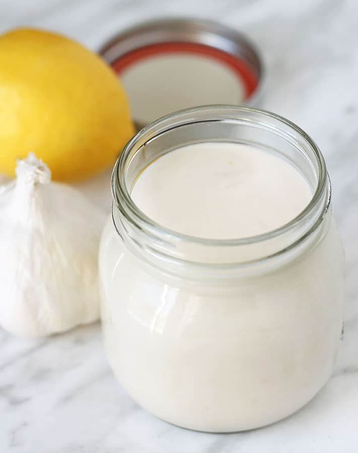 A jar of garlic aioli sauce sitting on a marble surface, a bulb of fresh garlic and a lemon sit to the left of the jar.