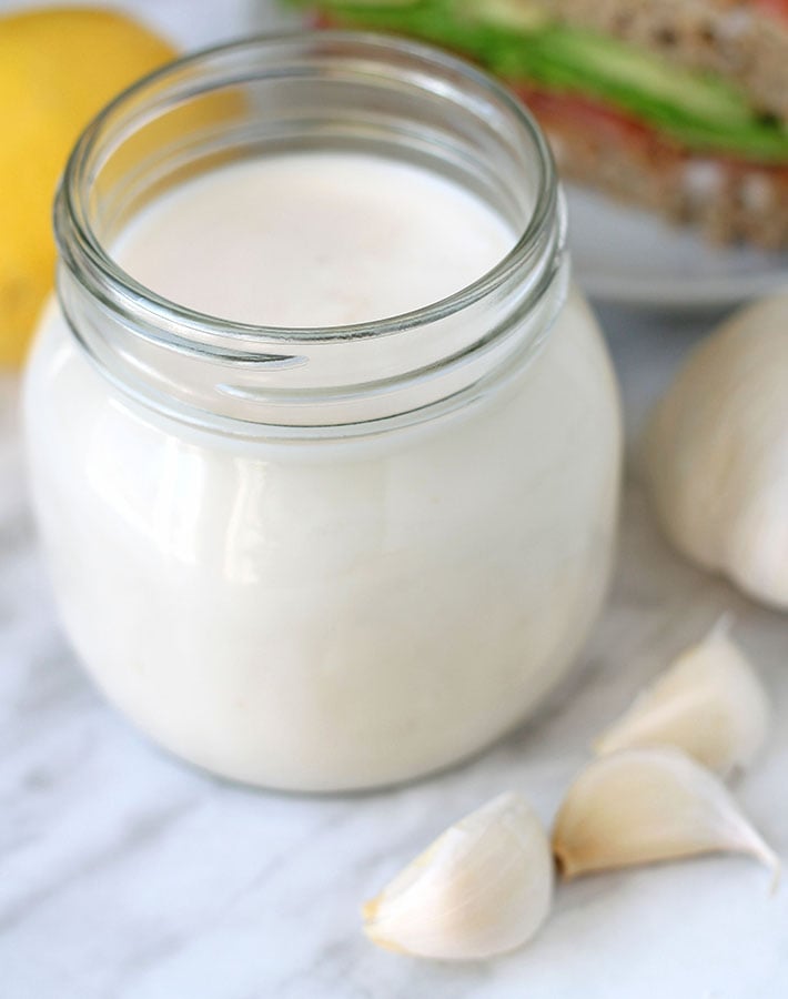 A glass mason jar with vegan aioli, three cloves of fresh garlic sit in front of the jar.