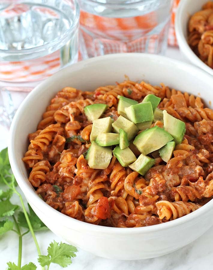 A bowl of One Pot Cheesy Taco Pasta on a marble surface, pasta is topped with cubed avocado.