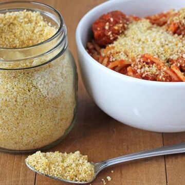Vegan Parmesan in a spoon on a wood table, a jar of it sits in the background as well as a bowl of spaghetti.