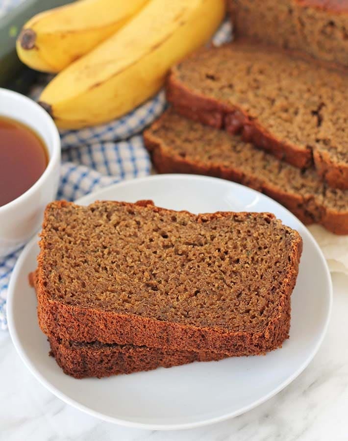 Two pieces of Gluten-Free Banana Zucchini Bread on a white plate, the rest of the loaf sits behind the plate.