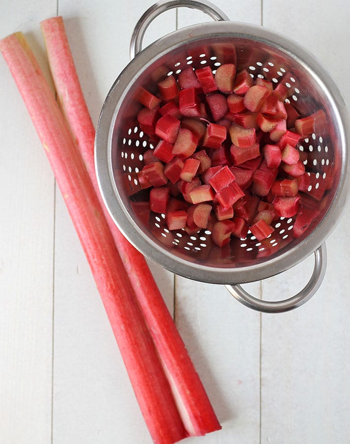 Two fresh rhubarb stalks on a white table, a small collander sits beside with chopped rhubarb.