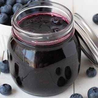 Small glass jar filled with vegan blueberry compote, jar is on a white table with fresh blueberries surrounding it.