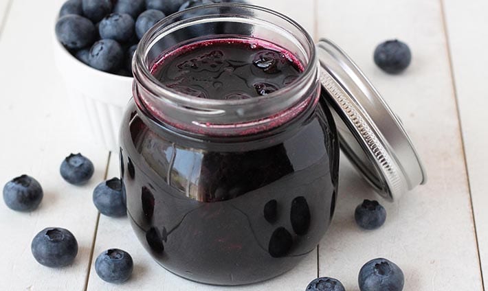 Small glass jar filled with vegan blueberry compote, jar is on a white table with fresh blueberries surrounding it.