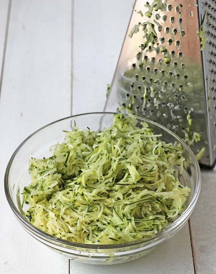 Shredded zucchini in a bowl with a greater behind the bowl.