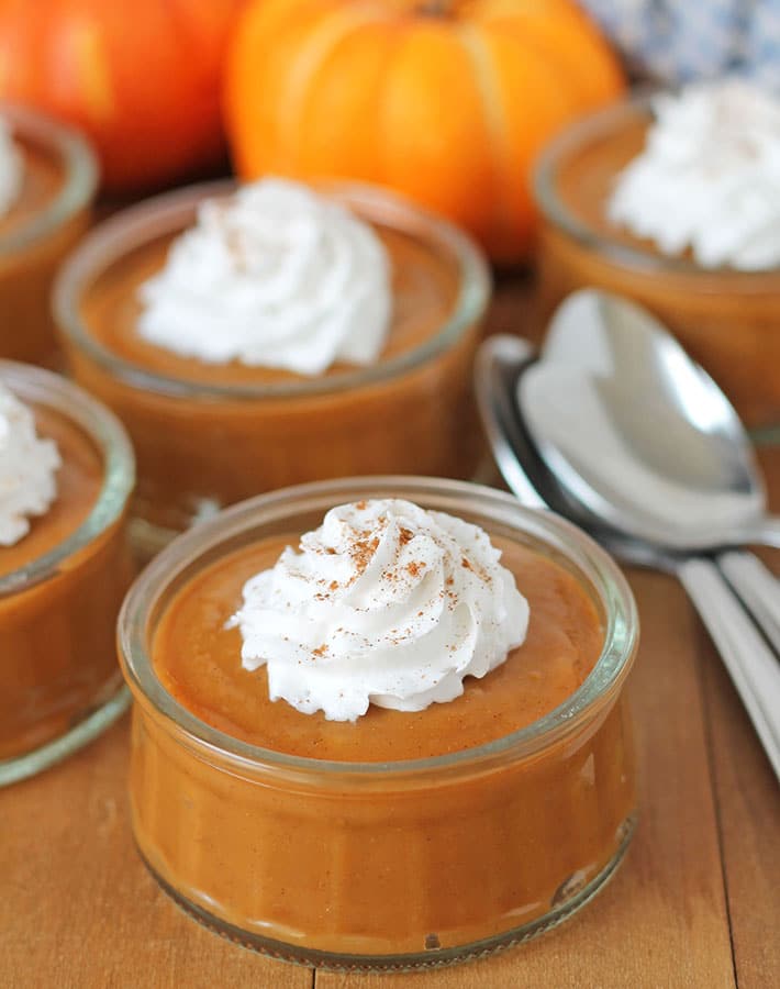 Vegan pumpkin pudding in glass cups on a brown wood table, pudding is topped with coconut whipped cream.