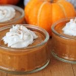 Vegan pumpkin pudding in glass serving jars sitting on a table, each pudding is topped with coconut cream.
