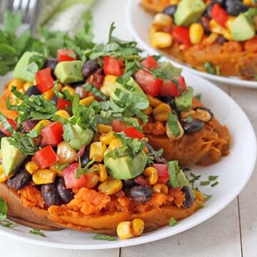 Two vegetarian stuffed sweet potatoes on a white plate, potatoes are garnished with avocado cubes and chopped cilantro.