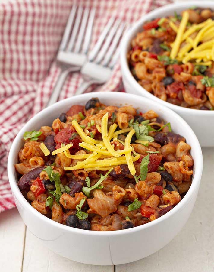 A bowl of vegetarian chili mac sitting on a table with two forks and another bowl behind it.