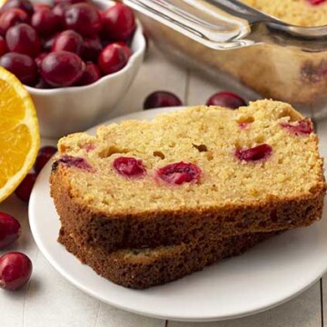 Two slices of gluten free cranberry orange bread on a plate, fresh canberries and oranges are around the plate.