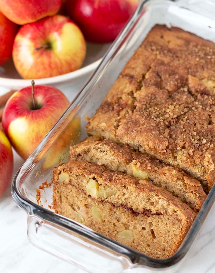 A loaf of apple bread in a glass baking dish, two pieces have been sliced and sit in front of the loaf.